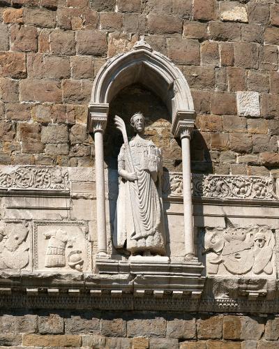 A close-up of a stone wall featuring a Gothic-style niche with a statue of a robed figure holding a palm frond, flanked by carved reliefs and set against a backdrop of aged masonry under a clear blue sky.
