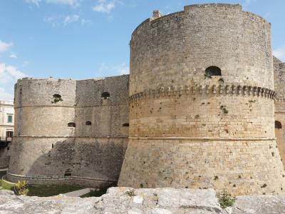 A low-angle view of a large, historic stone fortress featuring massive cylindrical towers with crenellations and arched windows, set against a blue sky with a modern building visible in the background.