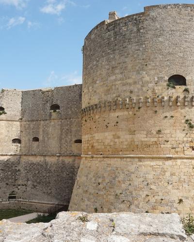 A low-angle view of a large, historic stone fortress featuring massive cylindrical towers with crenellations and arched windows, set against a blue sky with a modern building visible in the background.
