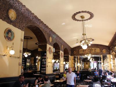 The image shows the interior of a historic café with distinctive Viennese Secession-style decor, including dark textured arches, ornate chandeliers, wall-mounted lamps, framed portraits, and patrons seated at tables.