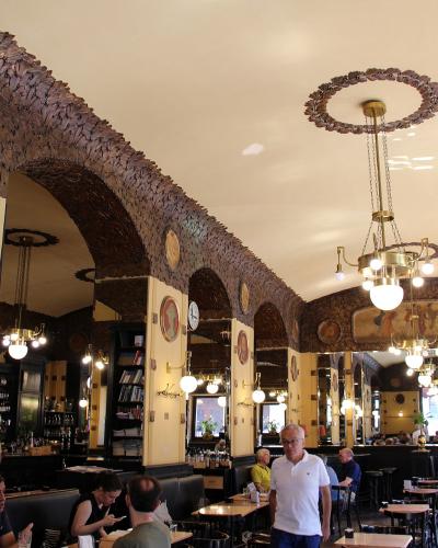 The image shows the interior of a historic café with distinctive Viennese Secession-style decor, including dark textured arches, ornate chandeliers, wall-mounted lamps, framed portraits, and patrons seated at tables.