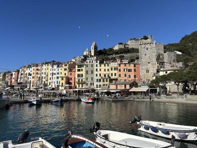 A harbor scene with colorful buildings along the waterfront, boats in the foreground, and a large stone castle tower on a hillside to the right under a clear blue sky.
