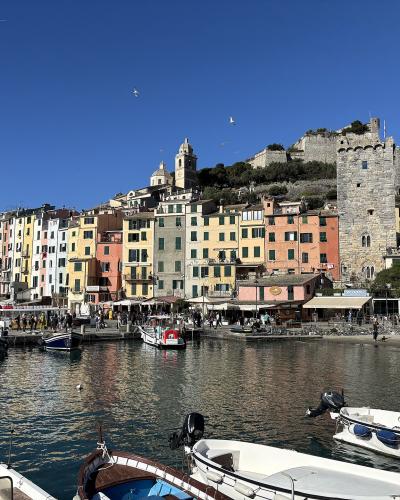 A harbor scene with colorful buildings along the waterfront, boats in the foreground, and a large stone castle tower on a hillside to the right under a clear blue sky.