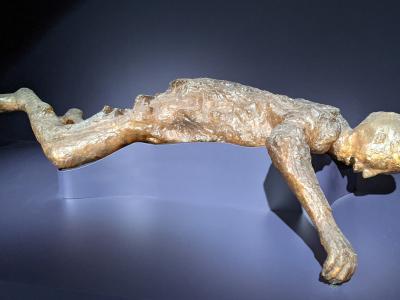 A plaster cast of a human victim from the eruption of Mount Vesuvius, displayed on a modern museum pedestal against a dark background.