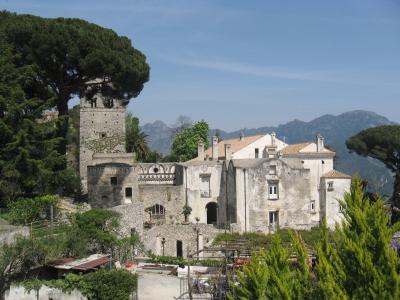 A stone medieval tower and adjacent whitewashed buildings with terracotta roofs, set on a hillside with lush greenery and mountains in the background under a clear blue sky.