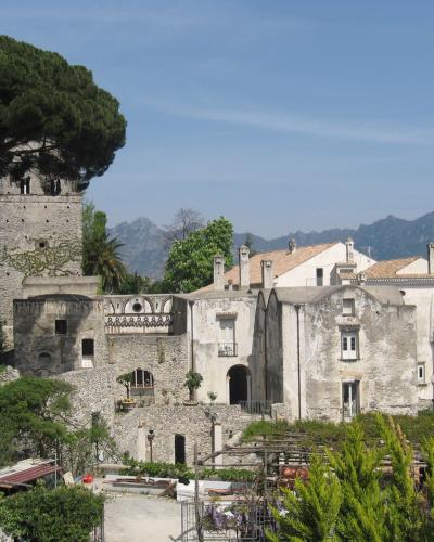A stone medieval tower and adjacent whitewashed buildings with terracotta roofs, set on a hillside with lush greenery and mountains in the background under a clear blue sky.