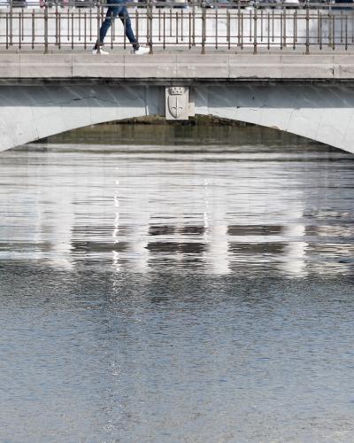 A low-angle view of a single-arch concrete bridge over a canal, with pedestrians walking on the deck and a stone coat of arms mounted on the arch's underside.