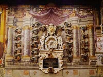 The image shows an ornate interior wall featuring a large, painted trompe-l'œil architectural facade with columns, draped fabric, and numerous coats of arms, centered around a bust portrait and a Latin inscription plaque.