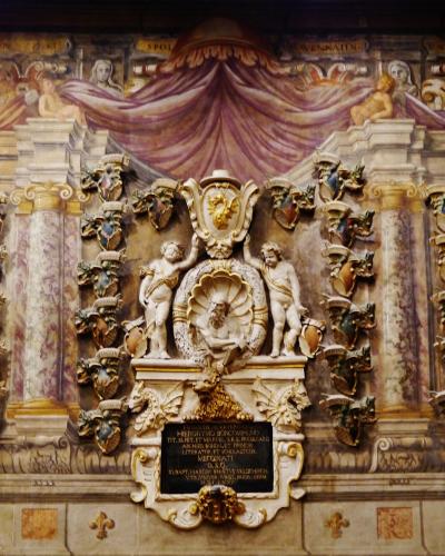 The image shows an ornate interior wall featuring a large, painted trompe-l'œil architectural facade with columns, draped fabric, and numerous coats of arms, centered around a bust portrait and a Latin inscription plaque.
