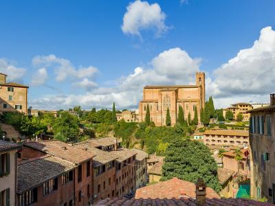 A sunlit view of Siena’s historic rooftops and terracotta buildings, with the large brick Gothic church of the Basilica of San Domenico prominently visible on a hill in the mid-ground under a blue sky with scattered clouds.