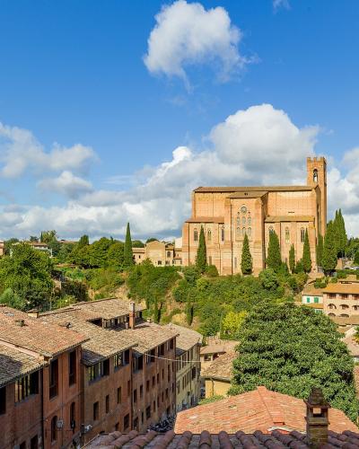 A sunlit view of Siena’s historic rooftops and terracotta buildings, with the large brick Gothic church of the Basilica of San Domenico prominently visible on a hill in the mid-ground under a blue sky with scattered clouds.
