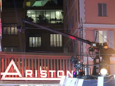 A nighttime exterior shot of a building facade featuring a large, illuminated neon sign reading "ARISTON" with a distinctive triangular logo, alongside professional television camera equipment on a crane and a cameraman operating it.
