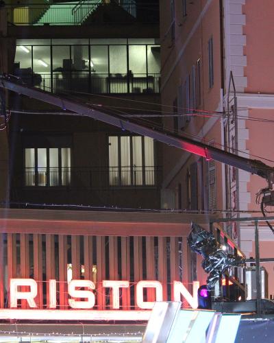 A nighttime exterior shot of a building facade featuring a large, illuminated neon sign reading "ARISTON" with a distinctive triangular logo, alongside professional television camera equipment on a crane and a cameraman operating it.