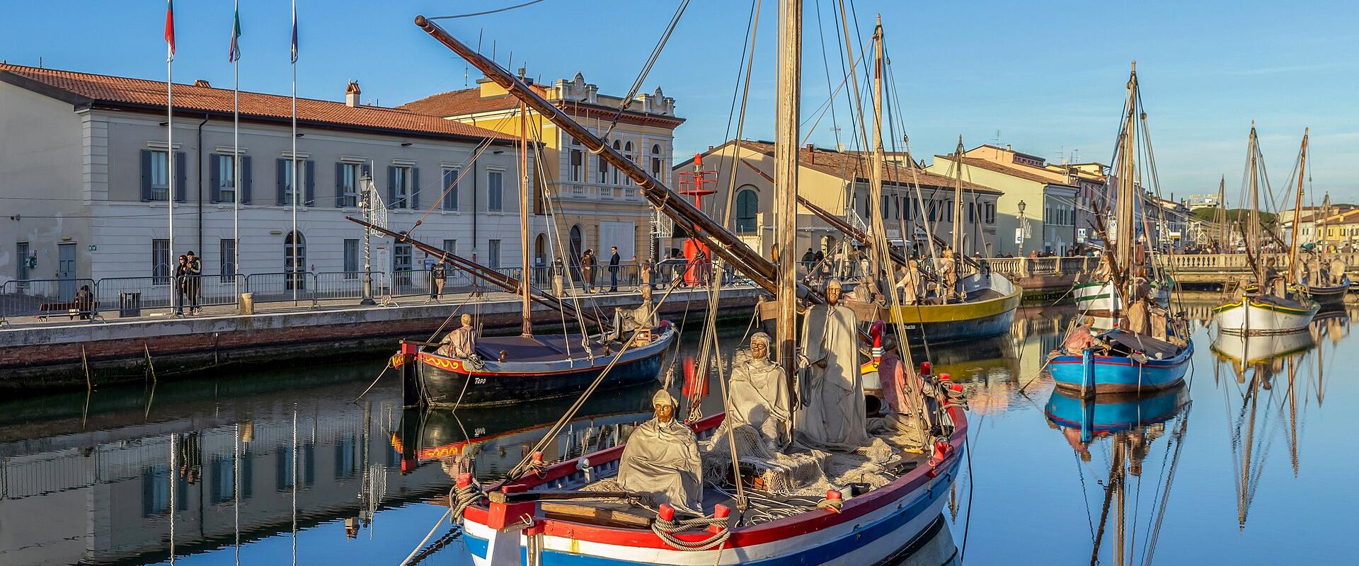 A canal scene in Cesenatico, Italy, featuring traditional wooden fishing boats (barche da pesca) with mannequins dressed as fishermen, moored along a historic waterfront lined with buildings and flags.