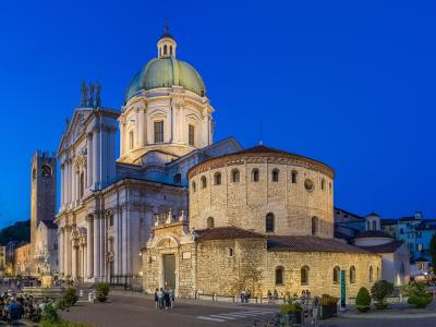 The image shows the Piazza del Duomo in Brescia, Italy, featuring the Duomo Nuovo (New Cathedral) with its prominent green dome and classical facade, alongside the Duomo Vecchio (Old Cathedral) with its Romanesque stone apse and bell tower, captur