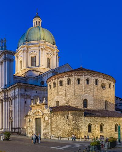 The image shows the Piazza del Duomo in Brescia, Italy, featuring the Duomo Nuovo (New Cathedral) with its prominent green dome and classical facade, alongside the Duomo Vecchio (Old Cathedral) with its Romanesque stone apse and bell tower, captur
