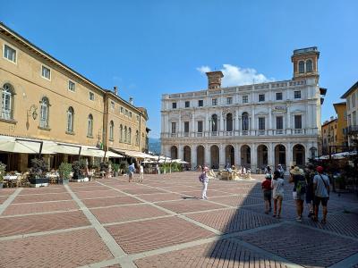 The image shows a large, historic stone building with Gothic arched windows and a central balcony, situated in a paved public square with a fountain featuring lion statues in the foreground and outdoor café seating to the right.