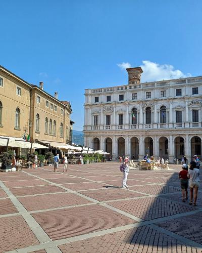 The image shows a large, historic stone building with Gothic arched windows and a central balcony, situated in a paved public square with a fountain featuring lion statues in the foreground and outdoor café seating to the right.