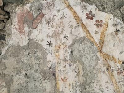 A close-up photograph of a weathered, peeling fresco on a rough stone wall, showing fragments of painted figures, stars, and floral motifs.
