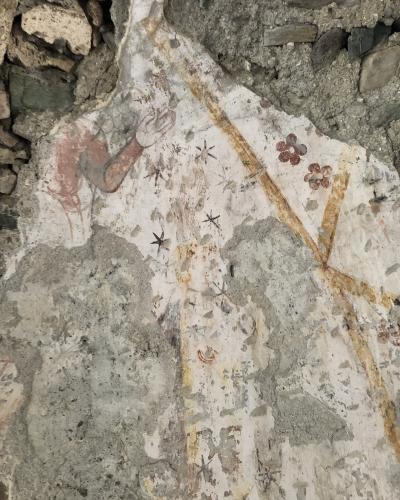 A close-up photograph of a weathered, peeling fresco on a rough stone wall, showing fragments of painted figures, stars, and floral motifs.