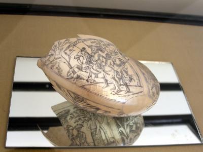 A close-up photograph of a nautilus shell intricately engraved with a scene of figures on horseback, displayed on a mirrored surface inside a glass case.