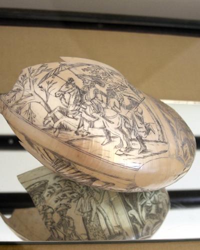 A close-up photograph of a nautilus shell intricately engraved with a scene of figures on horseback, displayed on a mirrored surface inside a glass case.