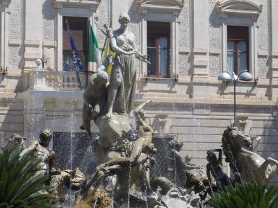 The image shows a large, ornate bronze fountain featuring a central female statue holding a bow and arrow, surrounded by other figures and water jets, set against a classical building facade with flags (EU, Italian, Sicilian) on a balcony.