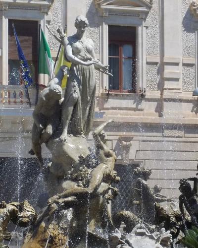 The image shows a large, ornate bronze fountain featuring a central female statue holding a bow and arrow, surrounded by other figures and water jets, set against a classical building facade with flags (EU, Italian, Sicilian) on a balcony.