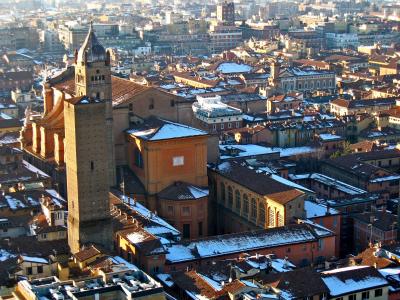 A high-angle view of the Basilica di San Pietro in Bologna, Italy, featuring its prominent brick bell tower (Torre di San Pietro) and adjacent church buildings, with snow on the rooftops and a cityscape in the background.