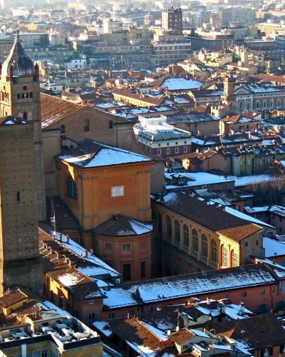 A high-angle view of the Basilica di San Pietro in Bologna, Italy, featuring its prominent brick bell tower (Torre di San Pietro) and adjacent church buildings, with snow on the rooftops and a cityscape in the background.