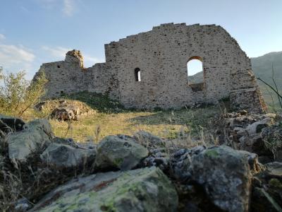 The image shows the stone ruins of a medieval castle or fortress, featuring a large arched window opening and crumbling walls, set against a backdrop of hills under a blue sky.
