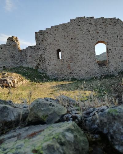 The image shows the stone ruins of a medieval castle or fortress, featuring a large arched window opening and crumbling walls, set against a backdrop of hills under a blue sky.