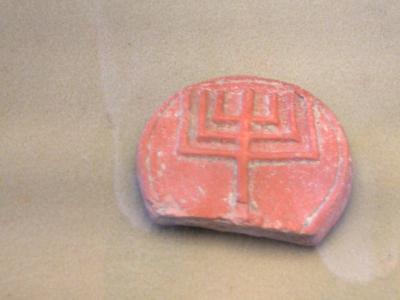 A close-up photograph of a reddish-orange, semi-circular clay seal impression featuring a geometric symbol resembling a trident or stylized plant, displayed inside a glass case.