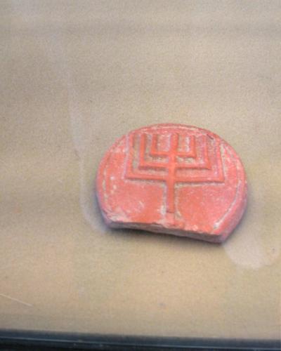 A close-up photograph of a reddish-orange, semi-circular clay seal impression featuring a geometric symbol resembling a trident or stylized plant, displayed inside a glass case.