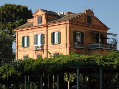 A two-story yellow-orange villa with green shutters, white balconies, and a tiled roof, surrounded by lush greenery and trees under a clear blue sky.