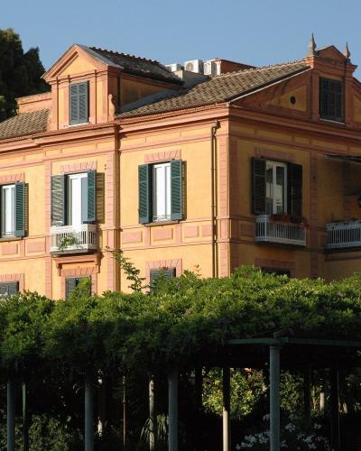 A two-story yellow-orange villa with green shutters, white balconies, and a tiled roof, surrounded by lush greenery and trees under a clear blue sky.