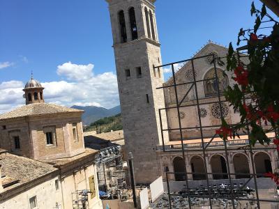 A sunlit Italian piazza with rows of white chairs set up for an event, framed by historic stone buildings including a tall bell tower and a cathedral facade with rose windows, under a blue sky with scattered clouds.