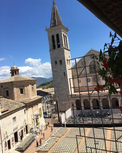A sunlit Italian piazza with rows of white chairs set up for an event, framed by historic stone buildings including a tall bell tower and a cathedral facade with rose windows, under a blue sky with scattered clouds.