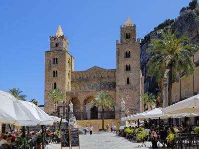 A large Norman-style cathedral with two prominent square towers and a central facade featuring arched windows and a rose window, situated in a paved public square with outdoor cafe seating under white umbrellas, palm trees, and a rocky cliff face