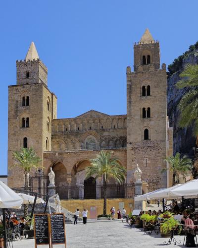 A large Norman-style cathedral with two prominent square towers and a central facade featuring arched windows and a rose window, situated in a paved public square with outdoor cafe seating under white umbrellas, palm trees, and a rocky cliff face