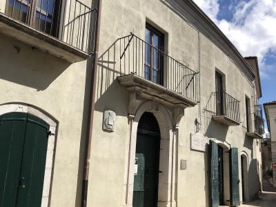 The image shows the exterior of a two-story building with a stone archway entrance, green wooden doors, and balconies with black metal railings under a blue sky; a plaque on the wall reads "PALAZZO DE LEO".