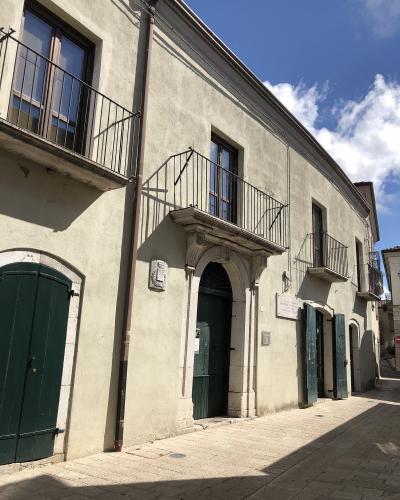 The image shows the exterior of a two-story building with a stone archway entrance, green wooden doors, and balconies with black metal railings under a blue sky; a plaque on the wall reads "PALAZZO DE LEO".