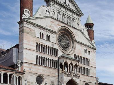 The image shows the facade of a large Romanesque cathedral with distinctive red brick towers capped with green copper spires, a prominent rose window, and a loggia at ground level, situated in a paved public square under a partly cloudy sky.