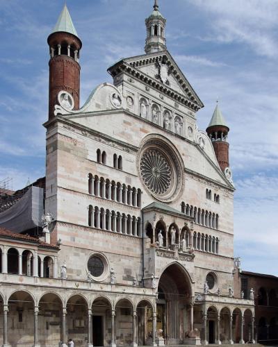 The image shows the facade of a large Romanesque cathedral with distinctive red brick towers capped with green copper spires, a prominent rose window, and a loggia at ground level, situated in a paved public square under a partly cloudy sky.