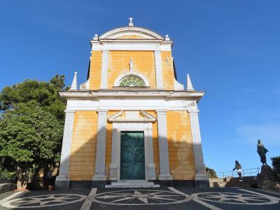 A low-angle shot of a yellow and white church facade with a green door, set against a blue sky, with a statue to the right and a mosaic compass rose on the ground in the foreground.