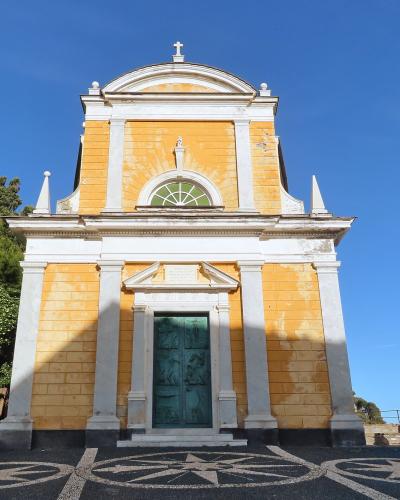 A low-angle shot of a yellow and white church facade with a green door, set against a blue sky, with a statue to the right and a mosaic compass rose on the ground in the foreground.