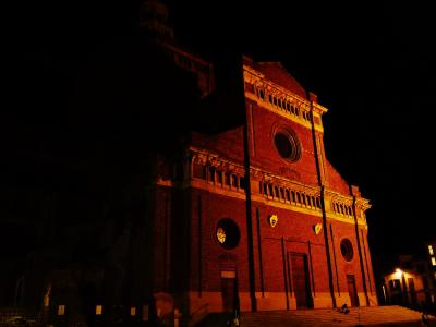 A low-angle night photograph of a large, illuminated red-brick cathedral facade featuring a central rose window, arched loggias, and prominent vertical buttresses, with steps leading up to the entrance.