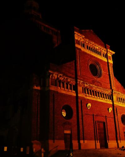 A low-angle night photograph of a large, illuminated red-brick cathedral facade featuring a central rose window, arched loggias, and prominent vertical buttresses, with steps leading up to the entrance.