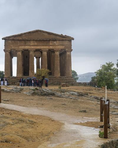 A panoramic view of the Valley of the Temples archaeological site in Agrigento, Sicily, featuring the Temple of Concordia prominently situated on a hilltop amidst olive groves and other ruins, with the modern city and coastline visible in the back