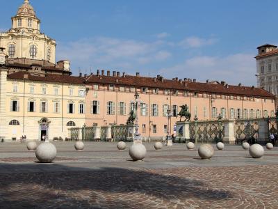 The image shows a large, paved public square with distinctive spherical stone bollards in the foreground, flanked by historic buildings including a prominent church with a dome on the left and a long pinkish palace behind an ornate iron gate featu